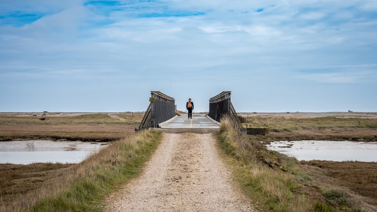 A man walking across the Bailey Bridge on Orford Ness with headphones on